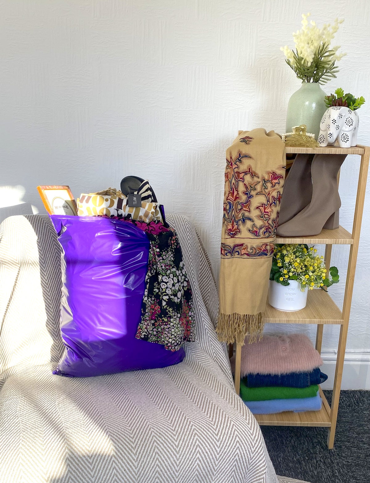 purple mailer bag filled with womens clothes which is sitting on a chair, next to wooden shelves which have clothes, bags, shoes and ornaments on it
