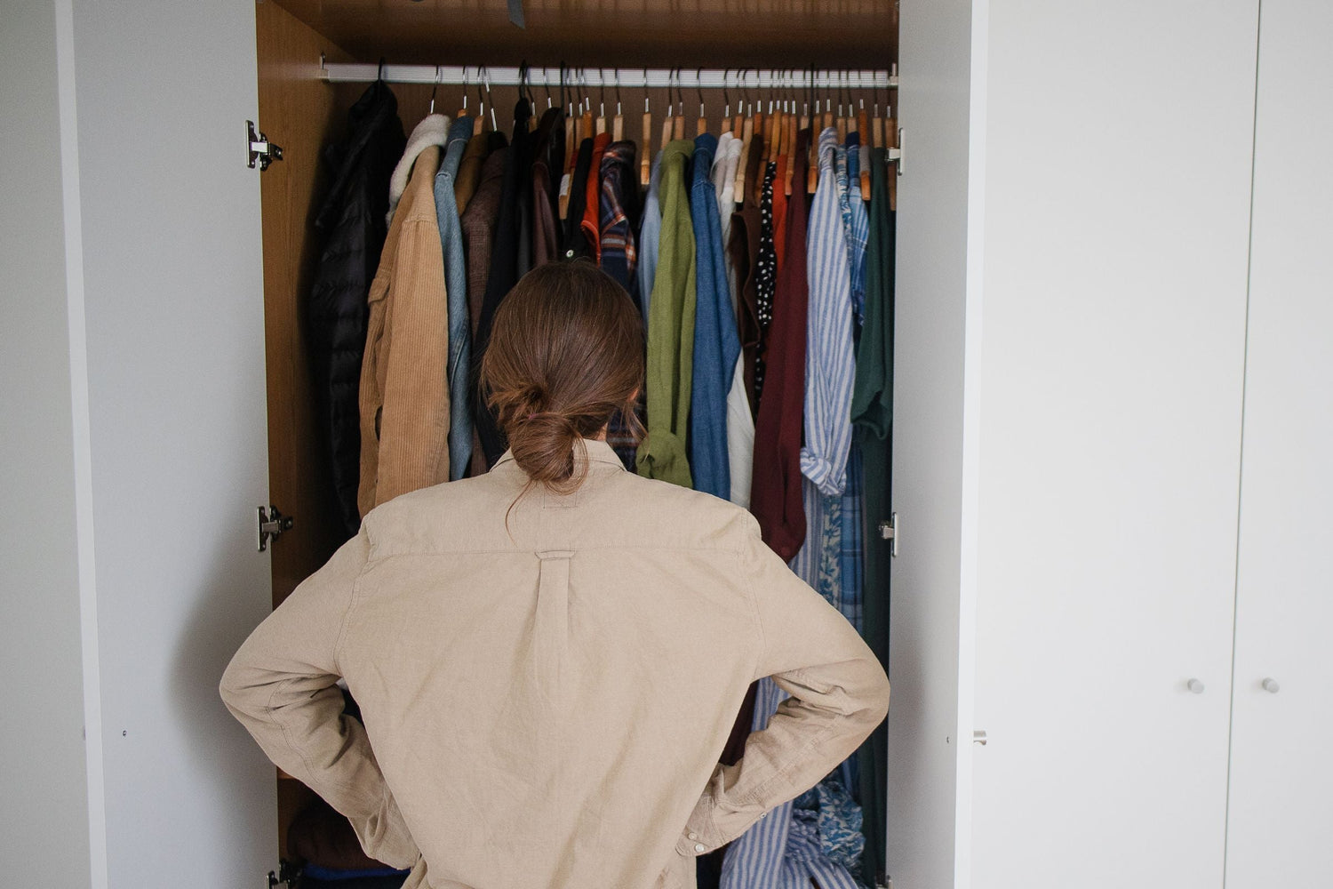 woman looking at a white wardrobe that is open and full of clothes, shot from behind