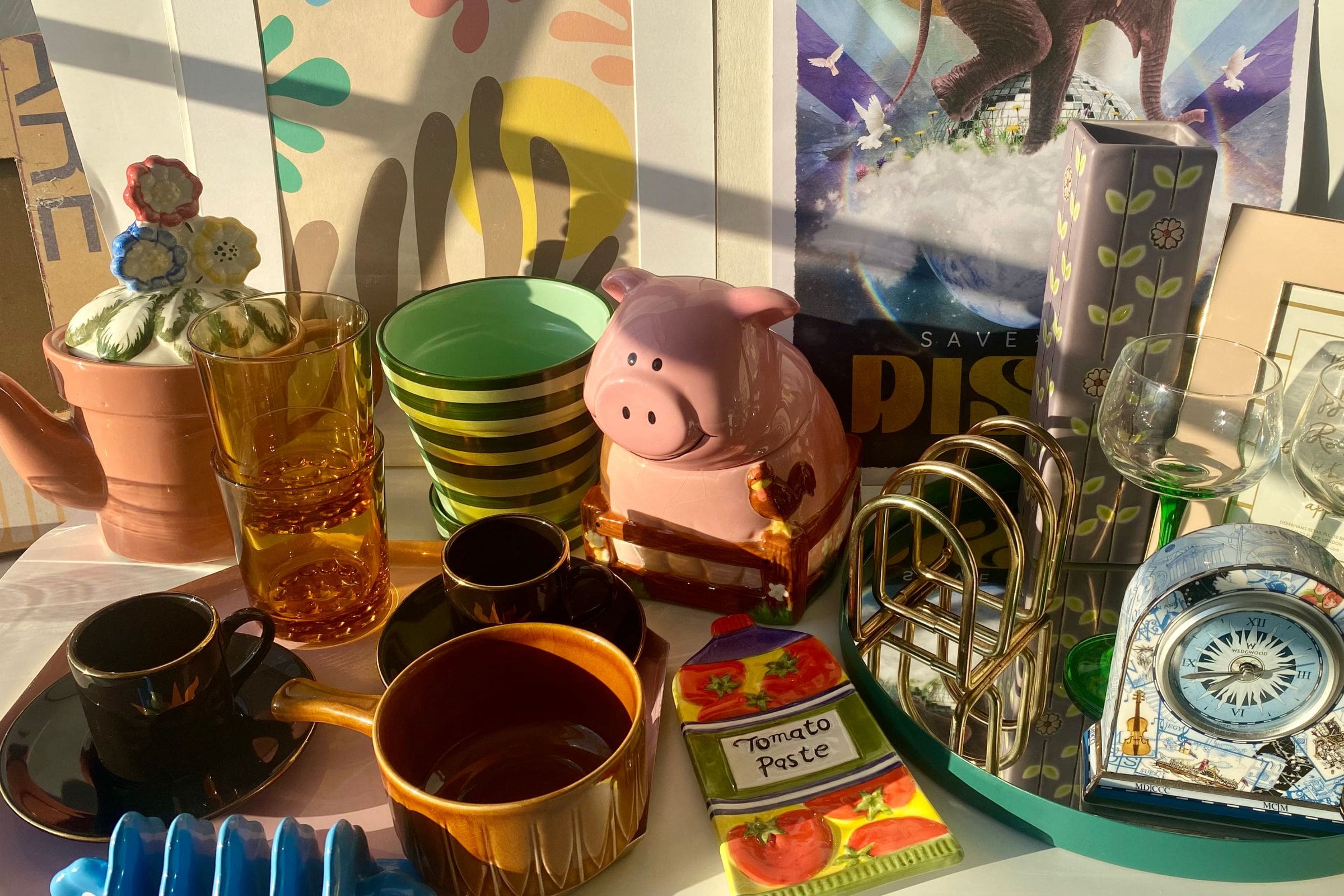 collection of unique home decor with the sun shining on them, the items include a pig biscuit tin, striped green plant pot and colourful print artwork