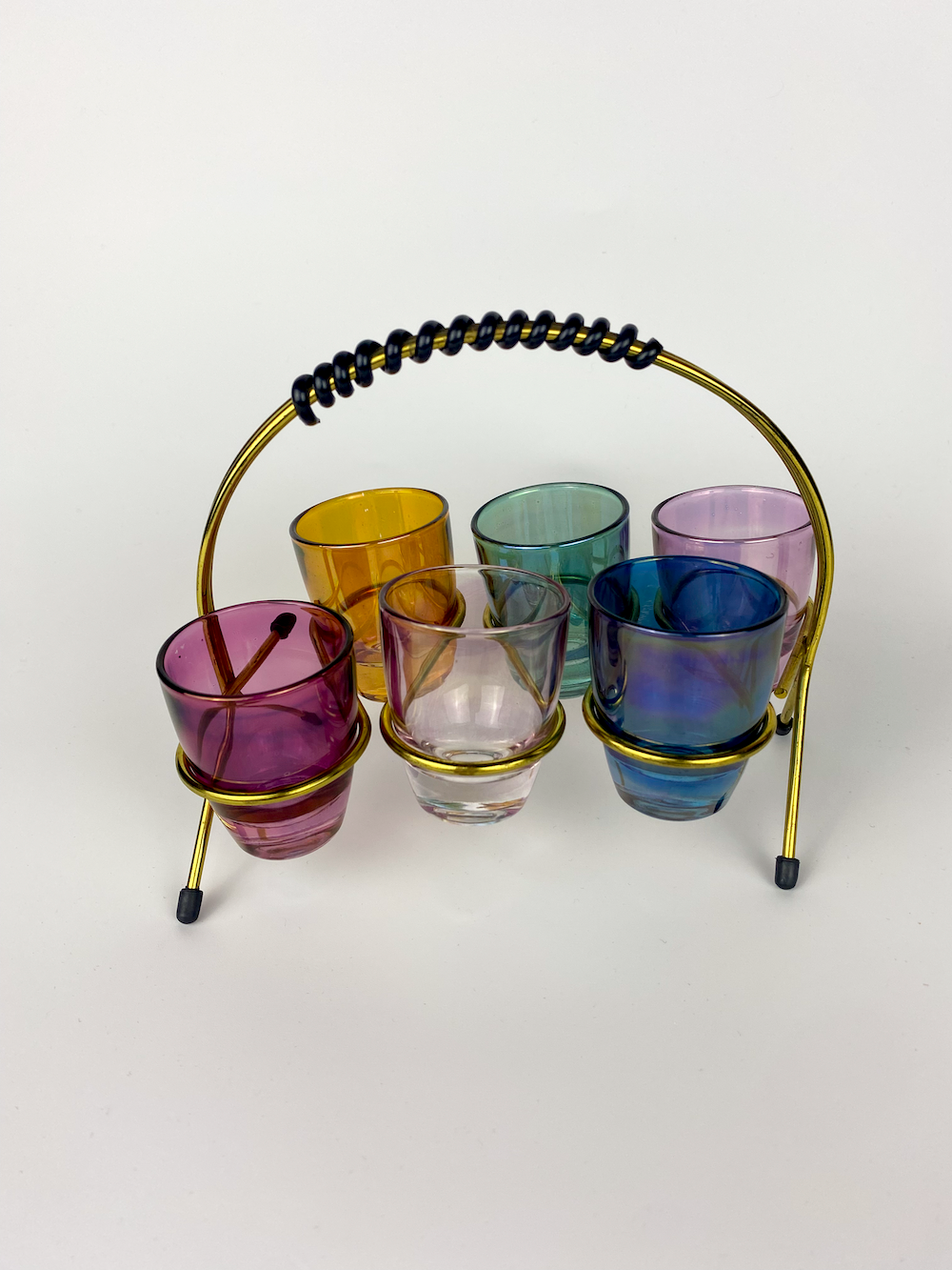 Multicoloured glass shot glasses sitting in a gold holder with a white background